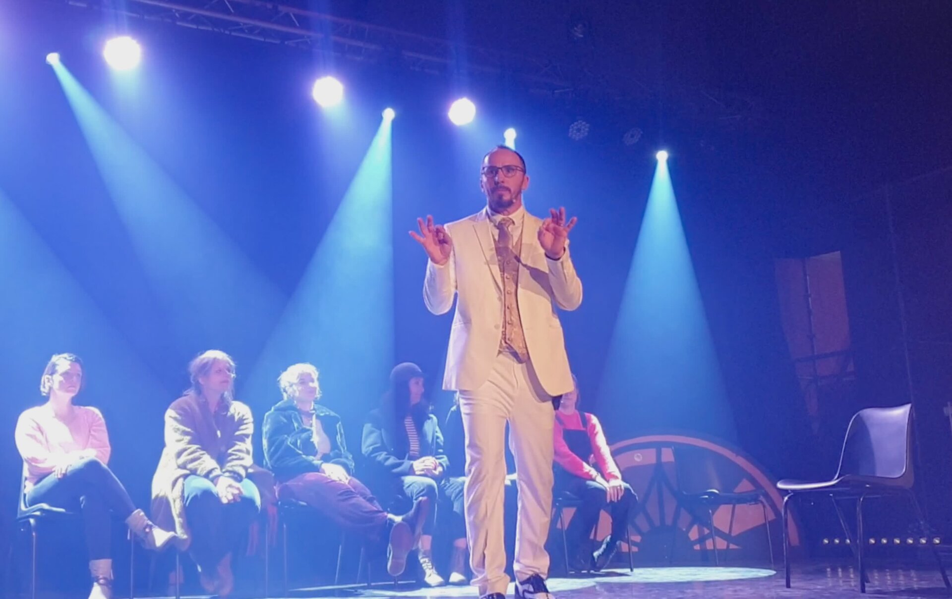Professeur M en costume clair, sur scène, projecteurs bleus